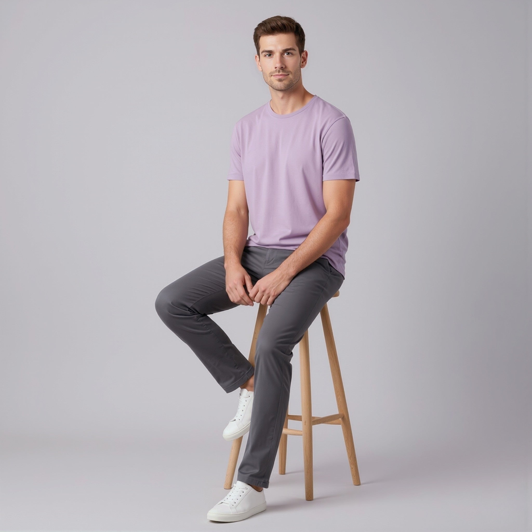 Man sitting on a wooden stool wearing a moleculuxe amethyst haze light purple t-shirt and gray pants against a plain background