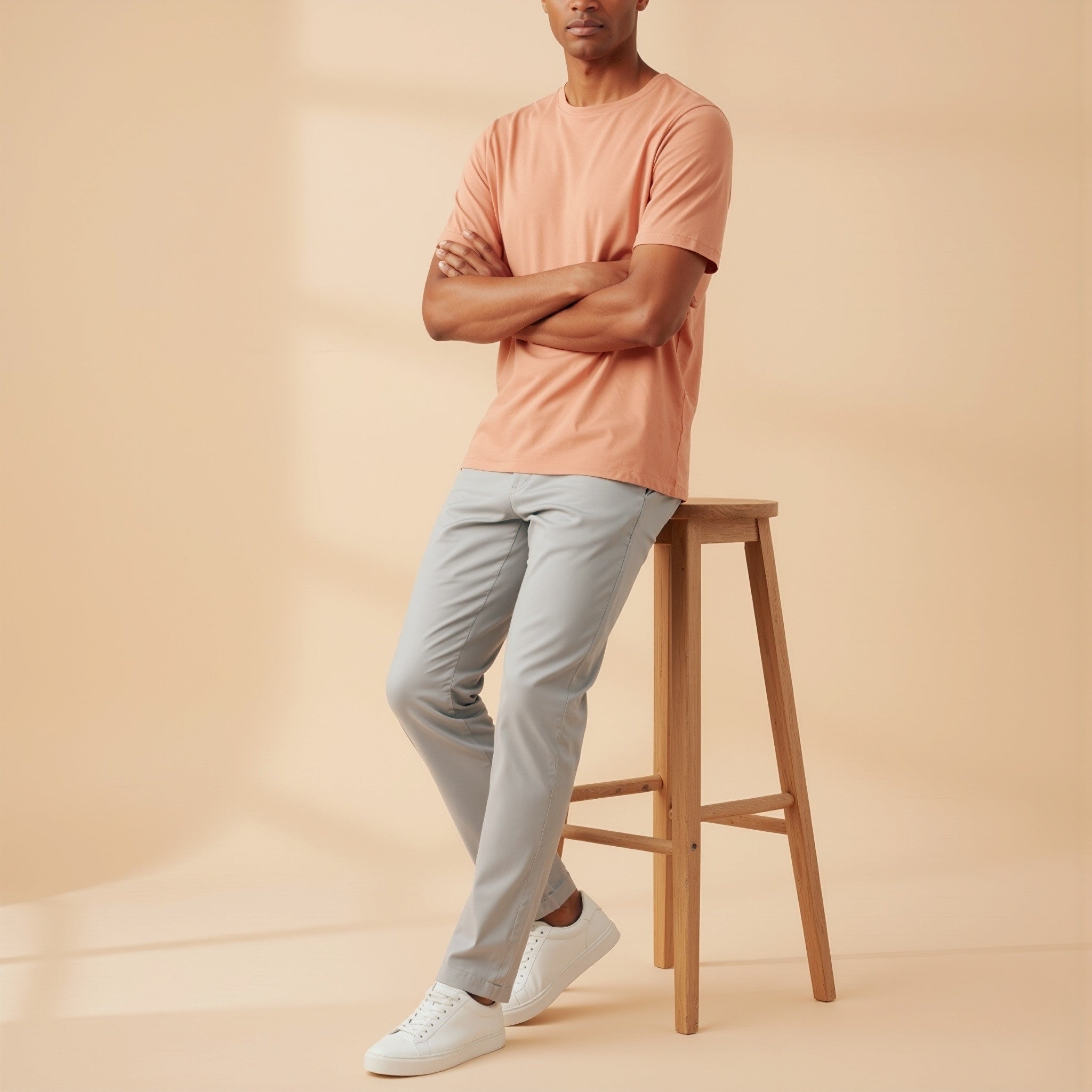 Man wearing a moleculuxe Apricot Silk  t-shirt and light gray pants standing on a wooden stool against a beige background