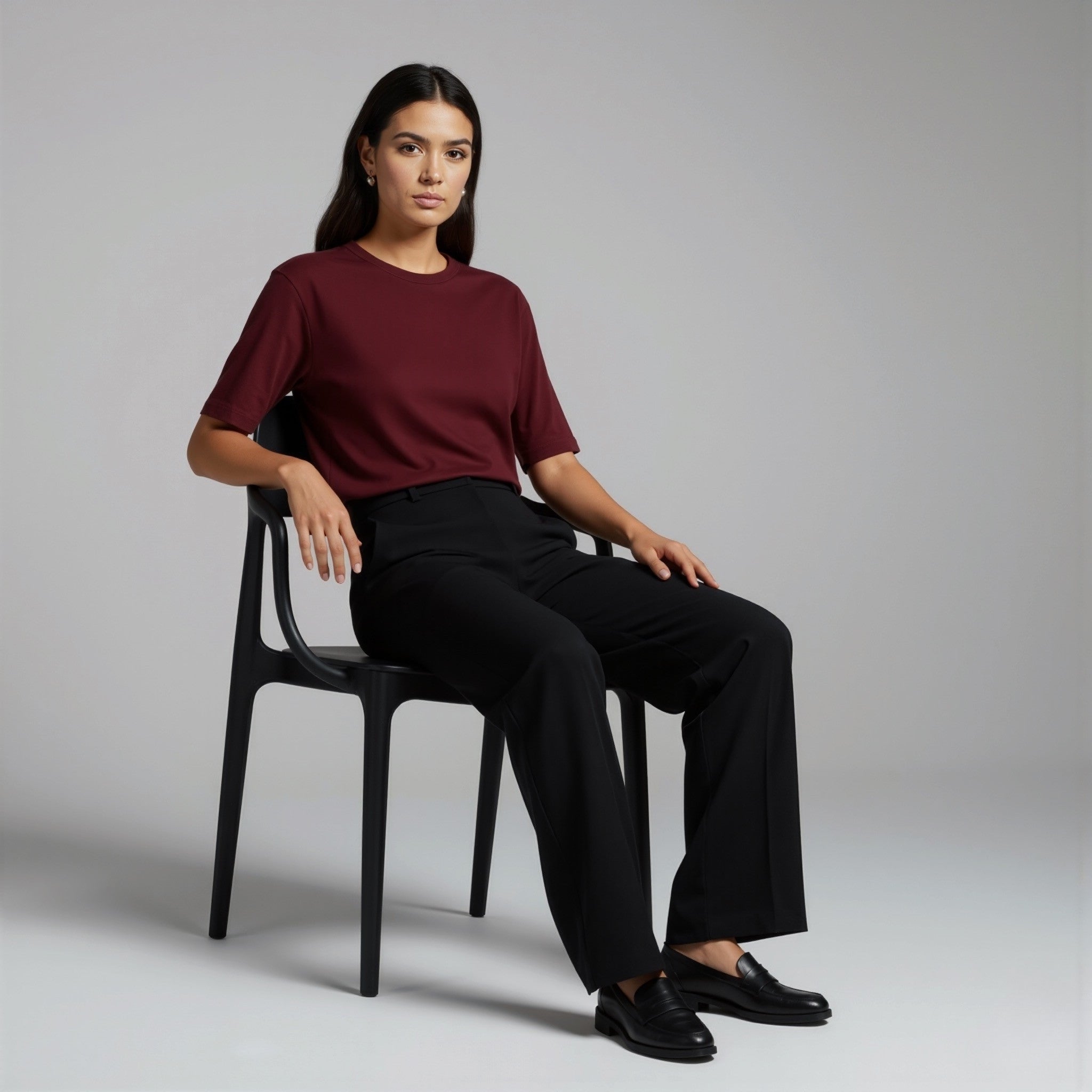 Woman sitting on a chair wearing a moleculuxe bordeaux burgundy top and black pants against a plain background