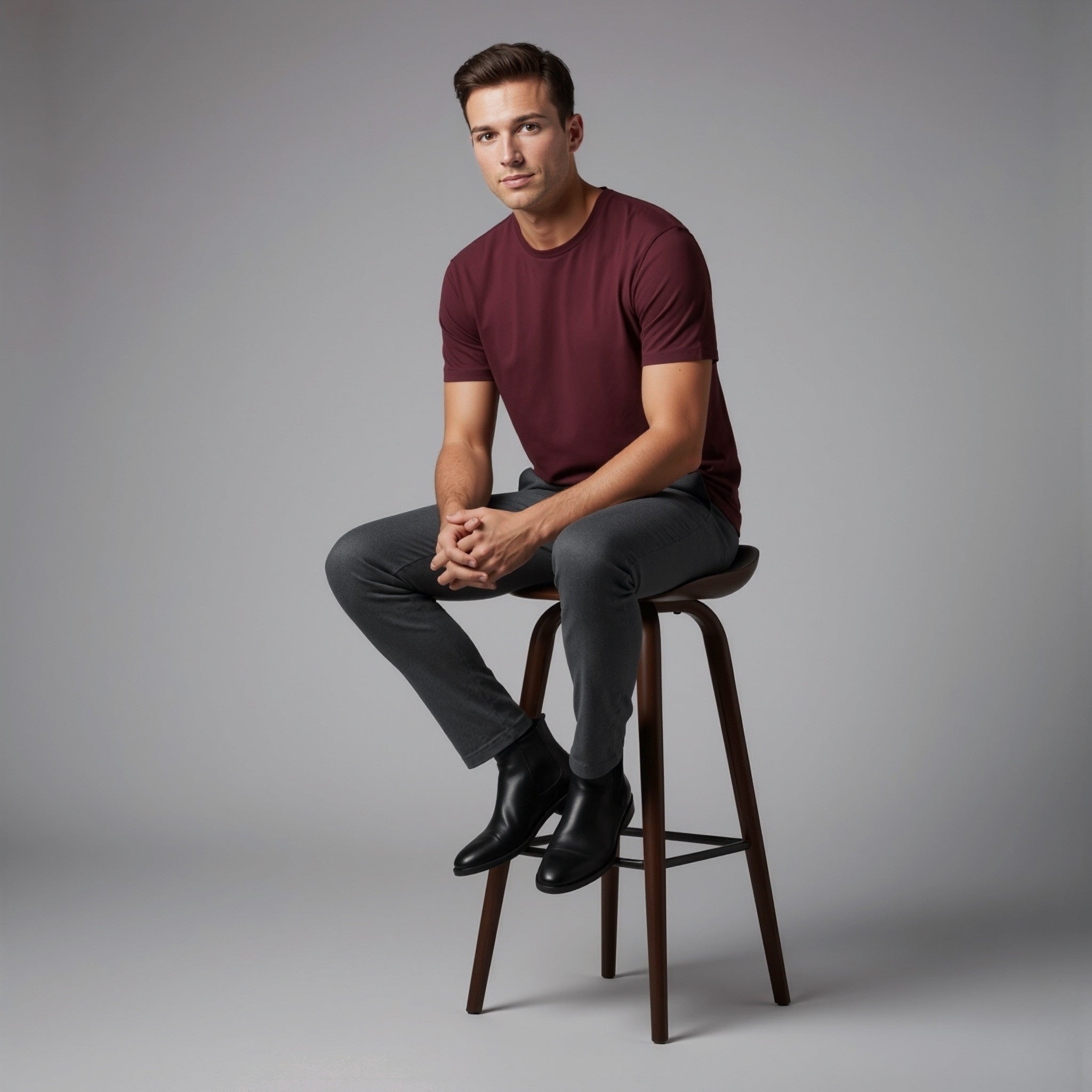 Man sitting on a stool wearing a moleculuxe bordeaux maroon shirt and gray pants against a gray background