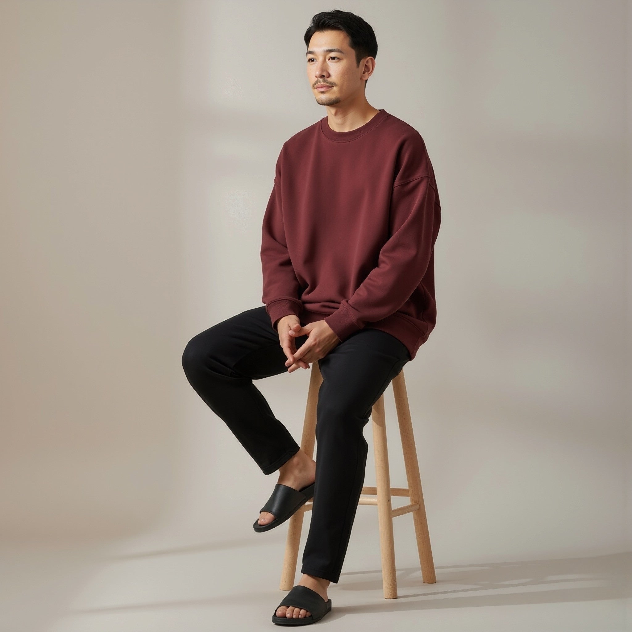 Man sitting on a wooden stool wearing a moleculuxe bordeaux maroon sweater and black pants against a plain background