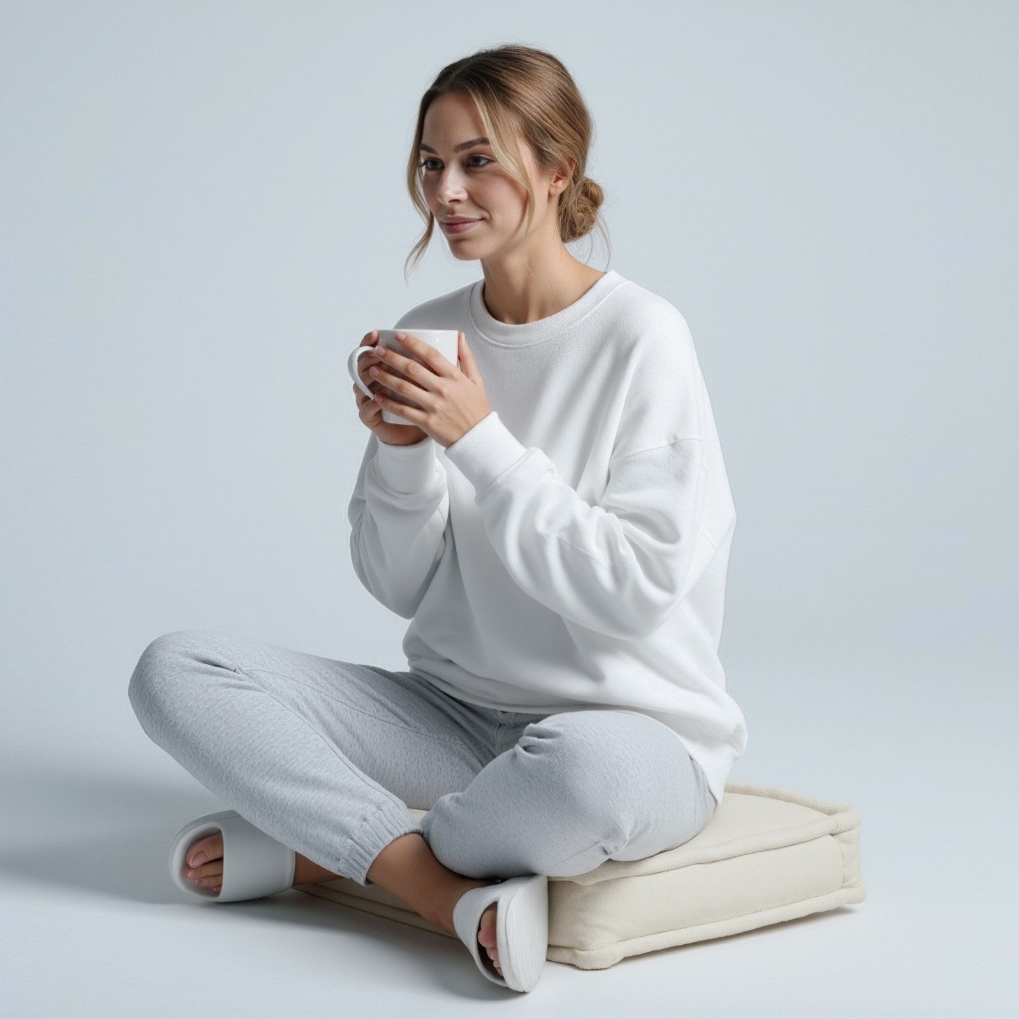 Woman sitting on a cushion wearing a moleculuxe pristine white sweatshirt and holding a mug against a light gray background
