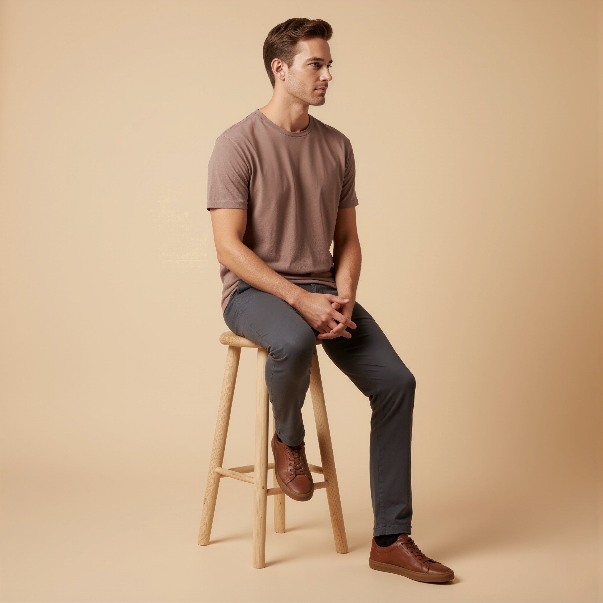 Man sitting on a wooden stool wearing a moleculuxe Truffle Taupe brown t-shirt and dark pants against a beige background