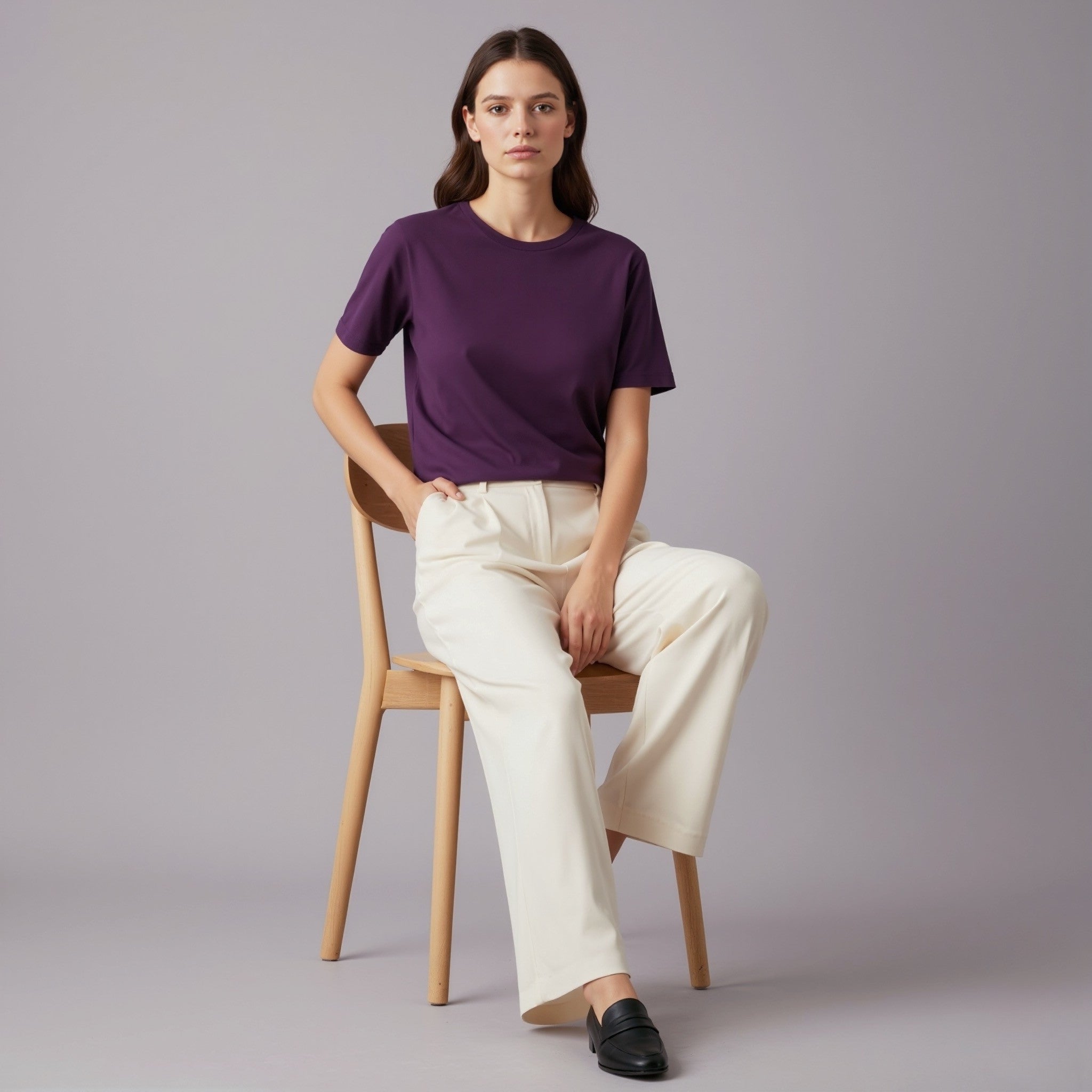 Woman sitting on a chair wearing a moleculuxe imperial violet purple shirt and white pants against a gray background