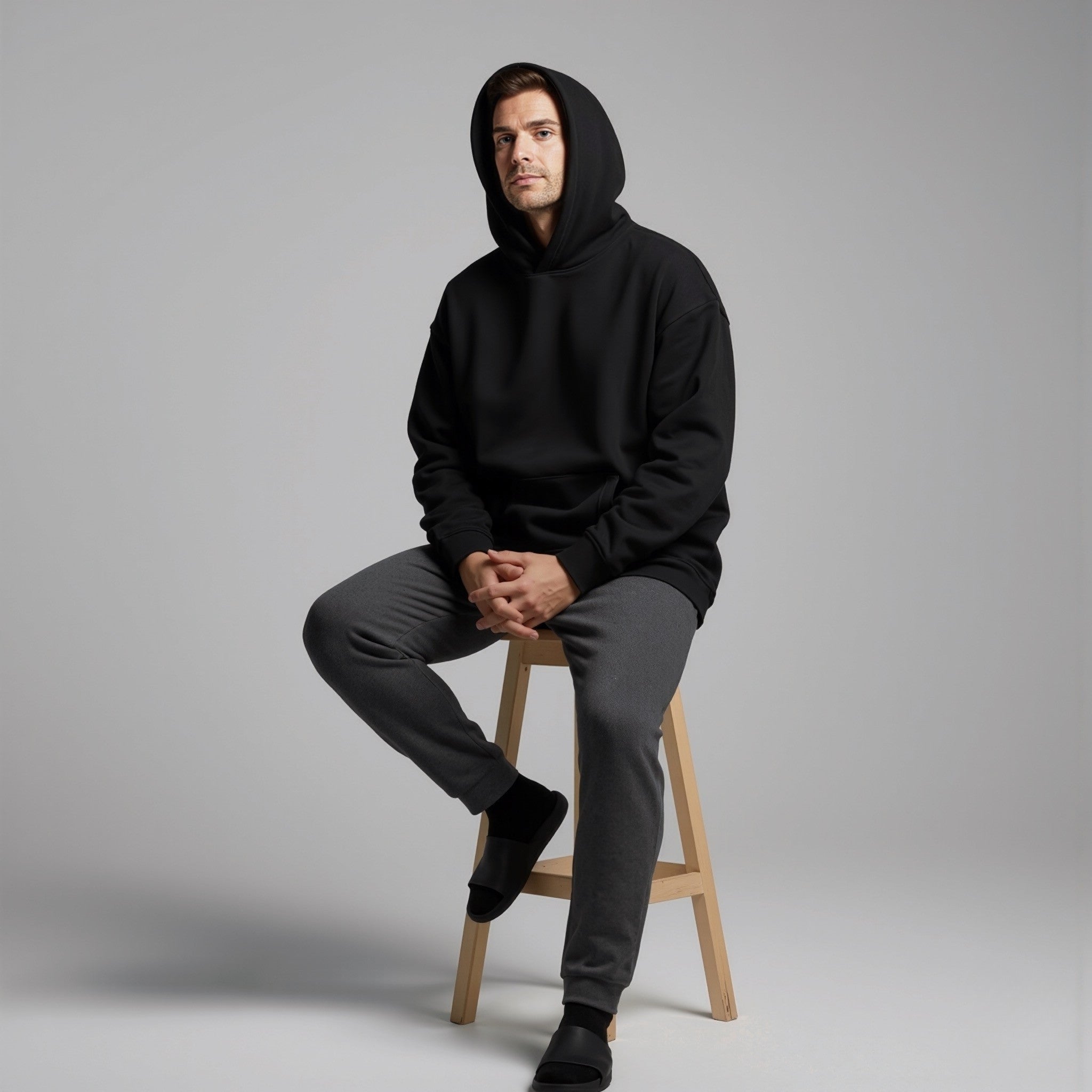 Man wearing a moleculuxe midnight noir black hoodie and gray pants sitting on a wooden stool against a gray background